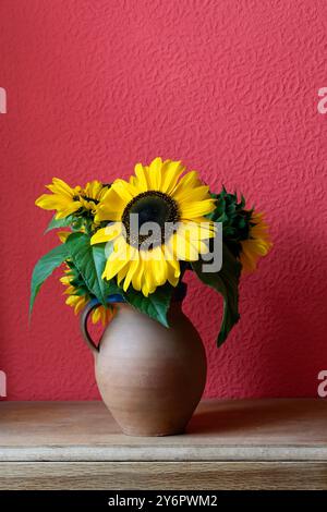 Ein Stillleben aus Sonnenblumen in einer Töpfervase vor einem rot strukturierten Hintergrund mit Kopierraum darüber Stockfoto