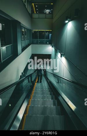 Ein Mann in einem Anzug, der eine Rolltreppe in einem modernen Bürogebäude mit reflektierenden Glaswänden und sanfter Beleuchtung herunterfährt. Stockfoto