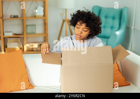 Afrikanisches Mädchen, das die Lieferung auspackt und in die Schachtel schaut. Glückliche Frau, die eine Schachtel öffnet. Eine Frau bekommt ein Paket, das sich die gelieferten Waren ansieht. Zufriedene Kunden positives Feedback, schnelle Expresslieferung Stockfoto