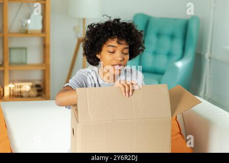 Afrikanisches Mädchen, das die Lieferung auspackt und in die Schachtel schaut. Glückliche Frau, die eine Schachtel öffnet. Eine Frau bekommt ein Paket, das sich die gelieferten Waren ansieht. Zufriedene Kunden positives Feedback, schnelle Expresslieferung Stockfoto
