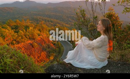 Eine ruhige Frau in weißem Kleid sitzt auf der Bergklippe mit Blick Stockfoto