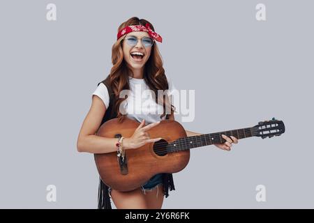 Attraktive, stilvolle junge Frau, die Akustikgitarre spielt und lächelt, während sie vor grauem Hintergrund steht Stockfoto