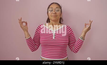 Eine heitere junge hispanische Frau meditiert an einer rosa Wand, ihre schöne Gelassenheit verkörpert Entspannung und Achtsamkeit. Stockfoto