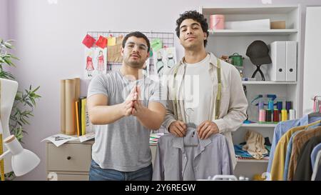 Zwei Schneiderinnen in einem mit Stoff gefüllten Atelier posieren mit entworfener Kleidung und zeigen ihr Handwerk. Stockfoto