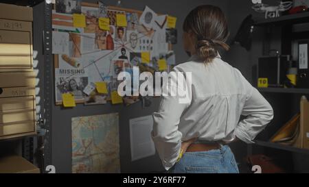 Eine junge Frau ermittelt in einem Detektivbüro, umgeben von Beweisstücken, Akten, Notizen und Karten. Stockfoto