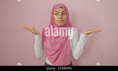 Eine junge Frau in einem rosafarbenen Hijab, die Ungewissheit vor einem isolierten rosa Hintergrund ausdrückt. Stockfoto