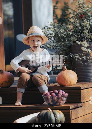 Ein kleiner Junge mit Hut, der eine Ukulele spielt, während er auf Holzstufen sitzt, umgeben von Kürbissen und Pflanzen. Stockfoto