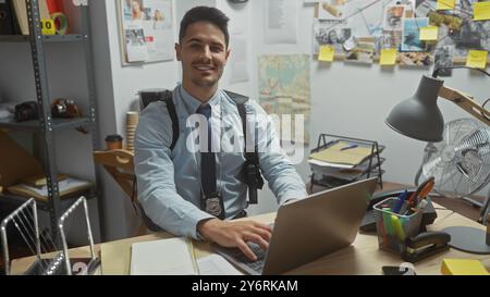 Ein selbstbewusster hispanischer Detective mit einem Ausweis in einem Büro, der an einem Laptop arbeitet und von Ermittlungsbeweisen umgeben ist. Stockfoto