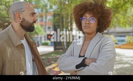 Mann und Frau in Business-Freizeitkleidung, die eine Diskussion in einem sonnigen Stadtpark mit verschwommenen Stadtgebäuden im Hintergrund führen. Stockfoto