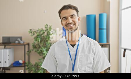 Porträt eines lächelnden jungen hispanischen männlichen Arztes, der einen Labormantel mit einem Lanyard in einer Reha-Klinik trägt. Stockfoto