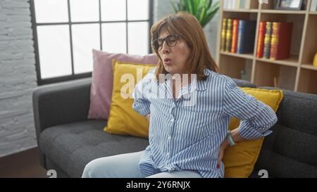 Frau mittleren Alters, die Rückenschmerzen erleidet, während sie auf einer Couch im Wohnzimmer sitzt und Beschwerden und Ängste zeigt. Stockfoto