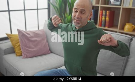 Ein kahler Mann dehnt sich in einem gemütlichen Wohnzimmer, trägt einen grünen Hoodie und bietet Komfort und Entspannung zu Hause. Stockfoto