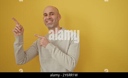 Lächelnder Glatzkopf mit Bart im Pullover, der vor gelbem Hintergrund nach oben zeigt. Stockfoto