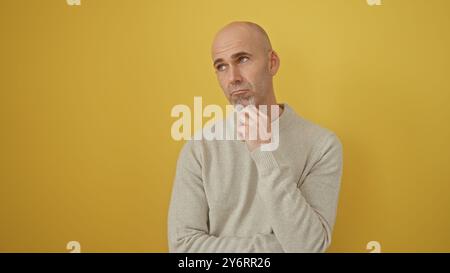 Nachdenklicher glatzhispanischer Mann mit Bart in grauem Pullover auf gelbem Hintergrund posiert nachdenklich. Stockfoto