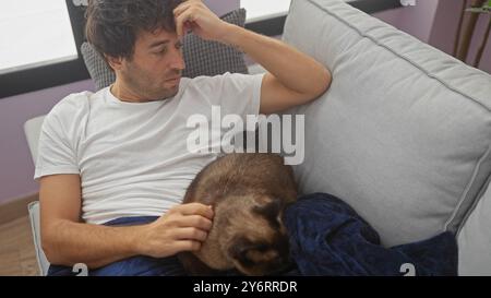 Ein junger hispanischer Mann entspannt sich mit seiner Katze auf einer Couch und verkörpert ungezwungenen Wohnkomfort. Stockfoto