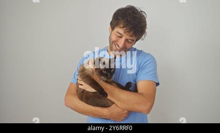 Ein glücklicher hispanischer Mann in einem blauen Hemd, der liebevoll eine siamesische Katze vor einem isolierten weißen Hintergrund hält Stockfoto