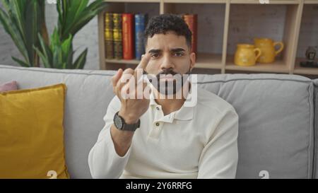 Ein junger hispanischer Mann gibt missbilligend mit seinem Mittelfinger, während er in einem gemütlichen Wohnzimmer sitzt. Stockfoto