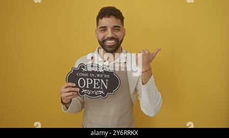 Hübscher bärtiger junger hispanischer Mann, der ein „offenes“ Schild hält und den Daumen seitlich über gelbem Hintergrund bewegt Stockfoto