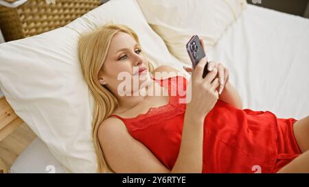 Eine junge blonde Frau in roter Loungewear benutzt ein Smartphone, während sie sich allein auf einem Bett in einem gut beleuchteten Schlafzimmer entspannt. Stockfoto