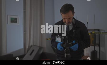 Ein reifer Mann mit Bart, Handschuhen und Ausweis untersucht eine Kamera an einem schwach beleuchteten Tatort. Stockfoto
