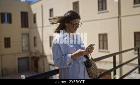 Eine junge hispanische Frau schlendert durch die alten Straßen von bari, italien, und schaut an einem sonnigen Tag in der historischen Region apulien aufmerksam auf ihr Handy Stockfoto