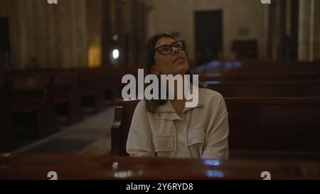 Eine junge hispanische Frau in einer Kirche in italien, die nach oben blickt, von Holzbänken umgeben und in sanftes Licht getaucht, in eine ruhige und reflektierende Atmosphäre getaucht Stockfoto