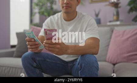 Ein junger Mann untersucht indische Währung, während er auf einer Couch in einem gemütlichen Wohnzimmer sitzt. Stockfoto