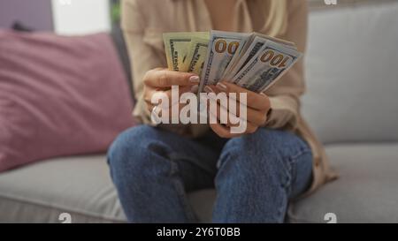 Eine junge Frau zählt uns Dollarscheine, während sie auf einer Couch in ihrem Wohnzimmer sitzt und eine Inneneinrichtung zeigt. Stockfoto