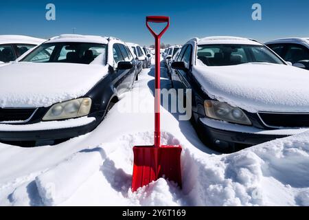 Autos werden nach einem schweren Schneesturm auf einem Parkplatz unter tiefem Schnee vergraben. Eine rote Schneeschaufel ragt in einer Schneewolke hervor. Der Prozess der Schneeräumung Stockfoto