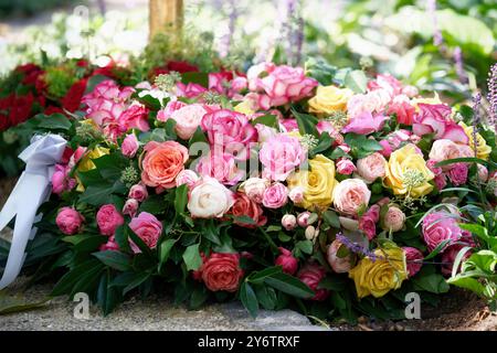 Ein Trauerkranz aus orangen, rosa, gelben und weißen Rosen und einem weißen Band liegt auf einem frischen Grab auf einem Friedhof Stockfoto