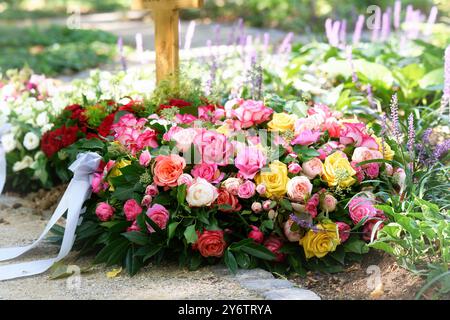 Ein Trauerkranz aus orangen, rosa, gelben und weißen Rosen und einem weißen Band liegt auf einem frischen Grab auf einem Friedhof Stockfoto