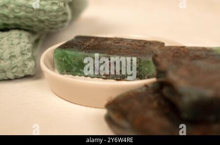 Zwei handgemachte Seifenriegel, ein grünes und ein braunes, sitzen auf einer weißen Schüssel. Ein grüner Waschlappen ist im Hintergrund unscharf. Stockfoto