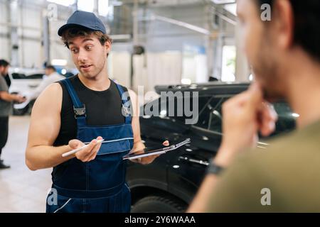 Fokussierter männlicher Garagenarbeiter in Uniform und nicht erkennbarer Mann Kunde spricht, um Details von Autoreparaturen zu besprechen. Stockfoto