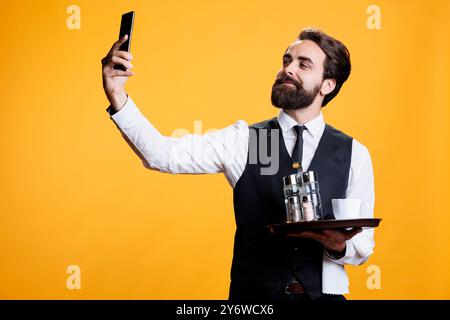 Lächelnde Person, die Fotos mit dem Telefon macht, arbeitet in der Gastronomie mit einem Tablett mit Besteck im Studio. Der professionelle Butler macht Fotos und Selfies in der Pause und macht Spaß mit dem Smartphone. Stockfoto