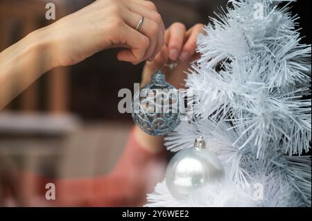 Blick durch ein Fenster mit weiblichen Händen, auf dem ein weißer Weihnachtsbaum mit einem glänzenden blauen weihnachtskugel hängt. Mit Reflexion des Außen in der gla Stockfoto
