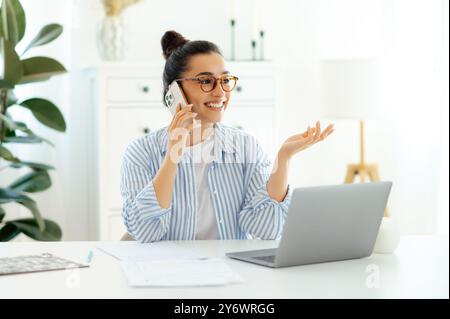 Mobiltelefongespräche. Eine selbstbewusste attraktive indische oder arabische Geschäftsfrau, die mit ihrem Smartphone mit einem Kunden oder Partner über die Strategie spricht, während sie tagtäglich im Büro mit einem Laptop auf dem Schreibtisch arbeitet, lächelt Stockfoto