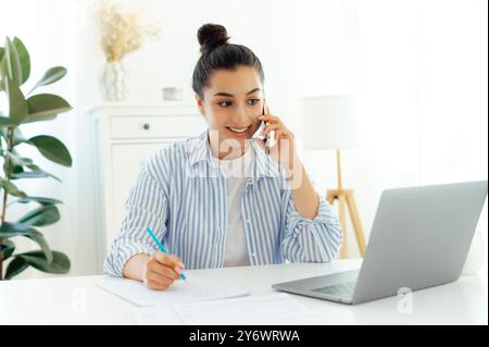 Smartphone-Konversation. Erfolgreiche junge, schöne weiße Frau mit Brille, die auf dem Handy und mit dem Laptop sitzt, während sie an ihrem Arbeitsplatz sitzt, mit einem Klienten über die Idee reden, lächeln Stockfoto