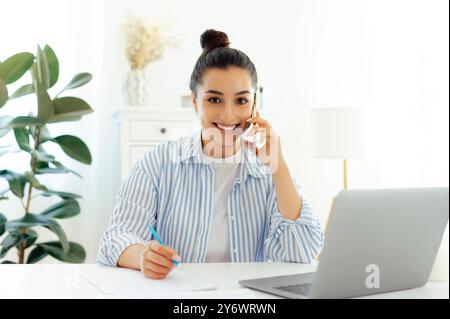 Telefongesprächskonzept. Erfolgreiche kaukasische Geschäftsfrau in blauem Hemd, die auf dem Mobiltelefon spricht und am Arbeitsplatz einen Laptop benutzt, mit dem Kunden ein Projekt bespricht und kamerafreundlich lächelt Stockfoto