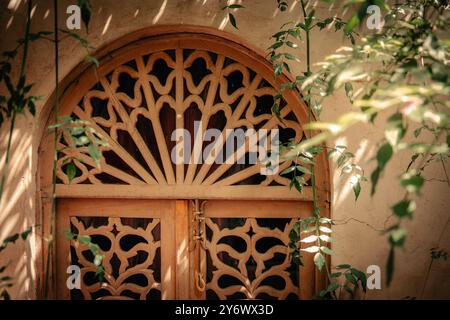 Kunstvolle marokkanische traditionelle Holztür mit kompliziertem Gitterdesign, strukturierter Wand und umliegendem Grün in einem ruhigen, luxuriösen Riad-Innenhof mit Stockfoto