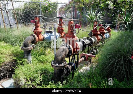 Installation von Industriewasserventilen und -Leitungen im Freien in einem landschaftlich gestalteten Bereich mit Gras und Sträuchern, gesichert durch einen Maschendrahtzaun im Hintergrund Stockfoto