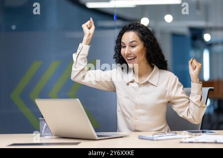 Geschäftsfrau, die ihre Begeisterung ausdrückt, während sie im modernen Büro an einem Laptop arbeitet. Happy Professional feiert Erfolge, was auf Leistung, Motivation und positive Emotionen hinweist. Stockfoto