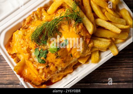 Nahaufnahme einer köstlichen Lasagne, garniert mit geschmolzenem Käse und garniert mit Kräutern, serviert mit knusprigen Pommes frites in einem Take-out-Behälter. Stockfoto