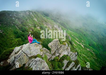 Der Wanderer sitzt am Rande eines großen Felsens mit Blick auf das üppige, grüne Tal mit steilen, felsigen Hängen. Frau, die sich einen Moment Zeit nimmt, um den Blick am Telefon einzufangen, umgeben von Nebel, der der Szene eine heitere, ätherische Qualität verleiht. Stockfoto