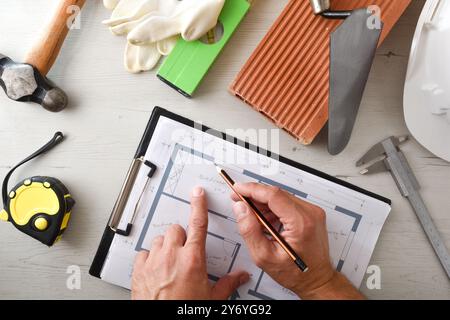 Architekt macht Notizen zum Hausplan auf weißem Holztisch mit Baumaterialien und Werkzeugen. Draufsicht. Stockfoto