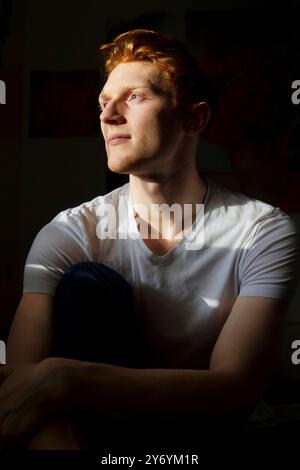 Ein junger Mann mit roten Haaren schaut nachdenklich aus einem sonnendurchfluteten Fenster Stockfoto