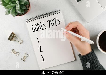 Frauenhand schreibt 2025 Tore in Notizbuch. Laptop, Hauspflanze, Tasse Kaffee auf Lichttisch. Draufsicht, flach Stockfoto