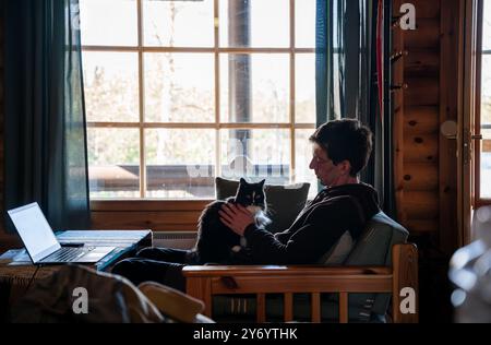 Mann, der auf dem Sofa sitzt und eine schwarze Katze vor einem großen Fenster hält Stockfoto