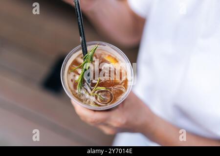 Blick von oben auf eine nicht erkennbare weibliche Hand, die am Sommertag eine Tasse Eiskaffee mit Stroh in der Stadt hält. Kaltes erfrischendes Getränk im Fokus. Stockfoto