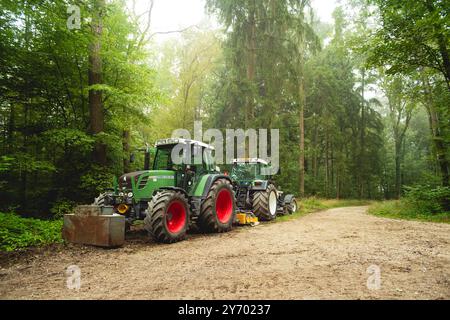 Große forstwirtschaftliche Traktoren arbeiten am Straßenrand in einem europäischen Wald. Nebliger Frühsommermorgen, weites, flaches Bild, keine Leute. Stockfoto