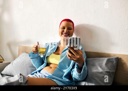 Eine junge Frau liegt auf ihrem Bett, lächelt, während sie einen Drink hält und ihr Telefon überprüft, und strahlt Freude aus. Stockfoto